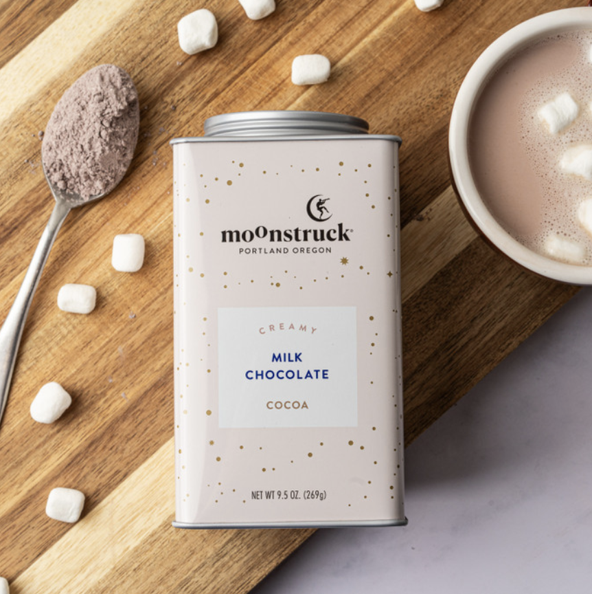 Moonstruck creamy milk chocolate cocoa canister on a wooden board with cocoa powder, marshmallows, and a cup of hot chocolate.
