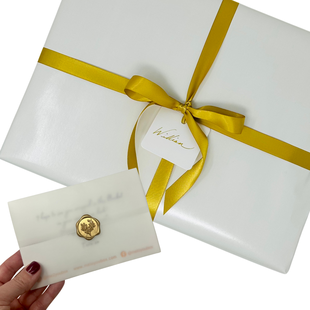 Gift wrapped box in white paper with gold satin ribbon. A hand with red nail polish is seen holding a gift message card next to the gift. The gift message card is 4x6 inches, wrapped in vellum and sealed with a gold wax seal.
