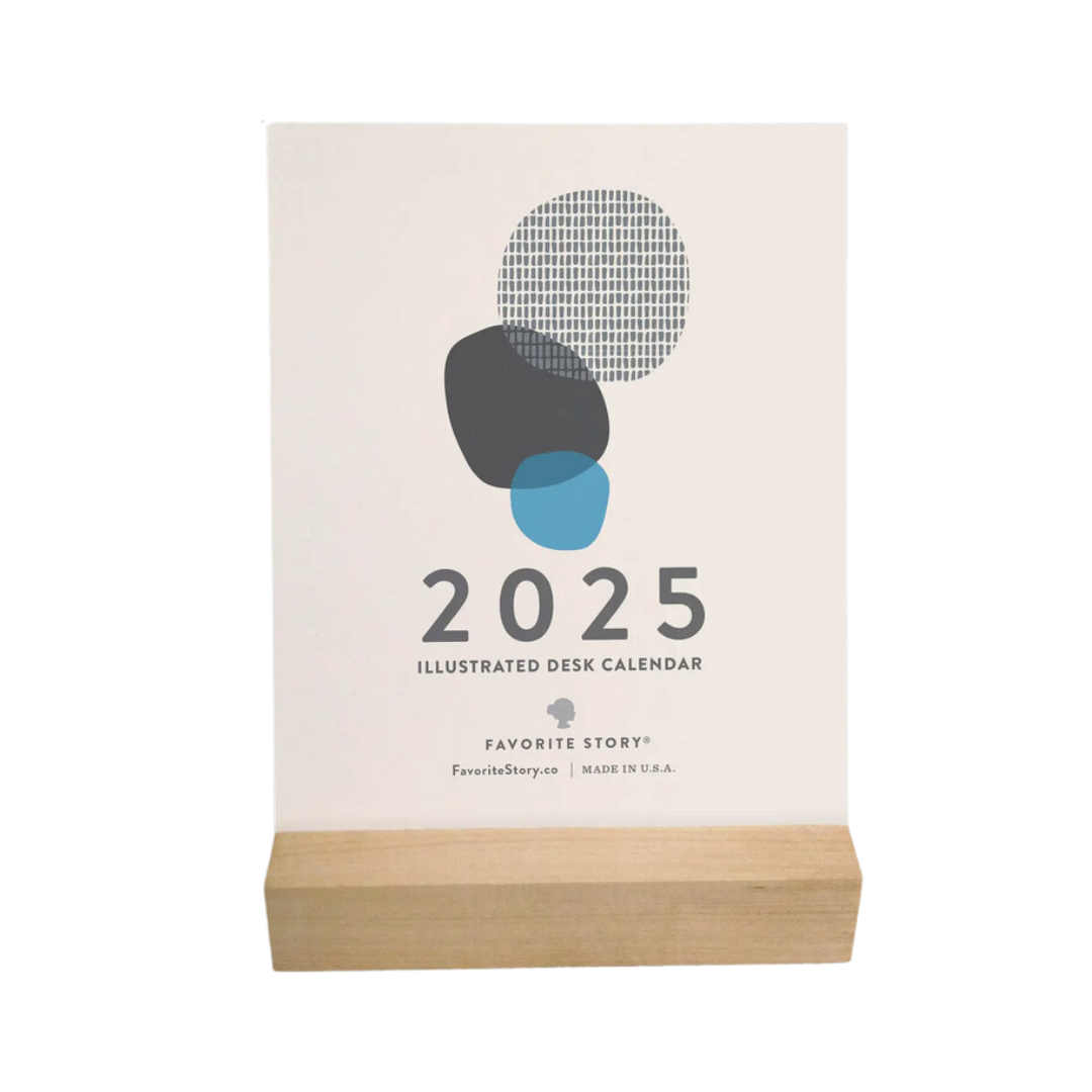 A 2025 Desk Calendar from Me To You Box displayed on a wooden desk. The calendar features a sleek maple wood stand and is set up with a monthly design card, showcasing vibrant artwork. The compact calendar size of 4.25 inches wide makes it fit neatly on the desk, highlighting its blend of style and functionality.