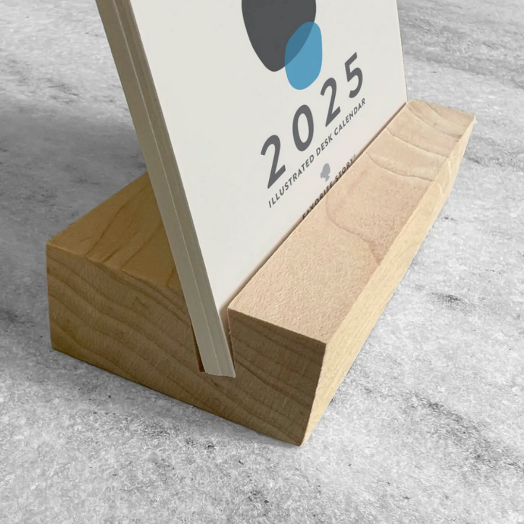 Close-up of the 2025 Desk Calendar with a unique monthly design visible on a 4.25" x 5.5" card. The calendar is positioned on a sturdy maple wood stand, emphasizing the quality craftsmanship. The calendar’s compact size is evident as it sits neatly on a small office desk.