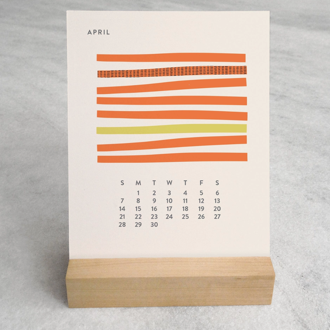 The 2025 Desk Calendar displayed on a table. The calendar is set on a desk with a focus on its maple wood stand and the monthly design cards. The compact size and elegant presentation are showcased, making it clear that this calendar is both a functional and stylish addition to any workspace.