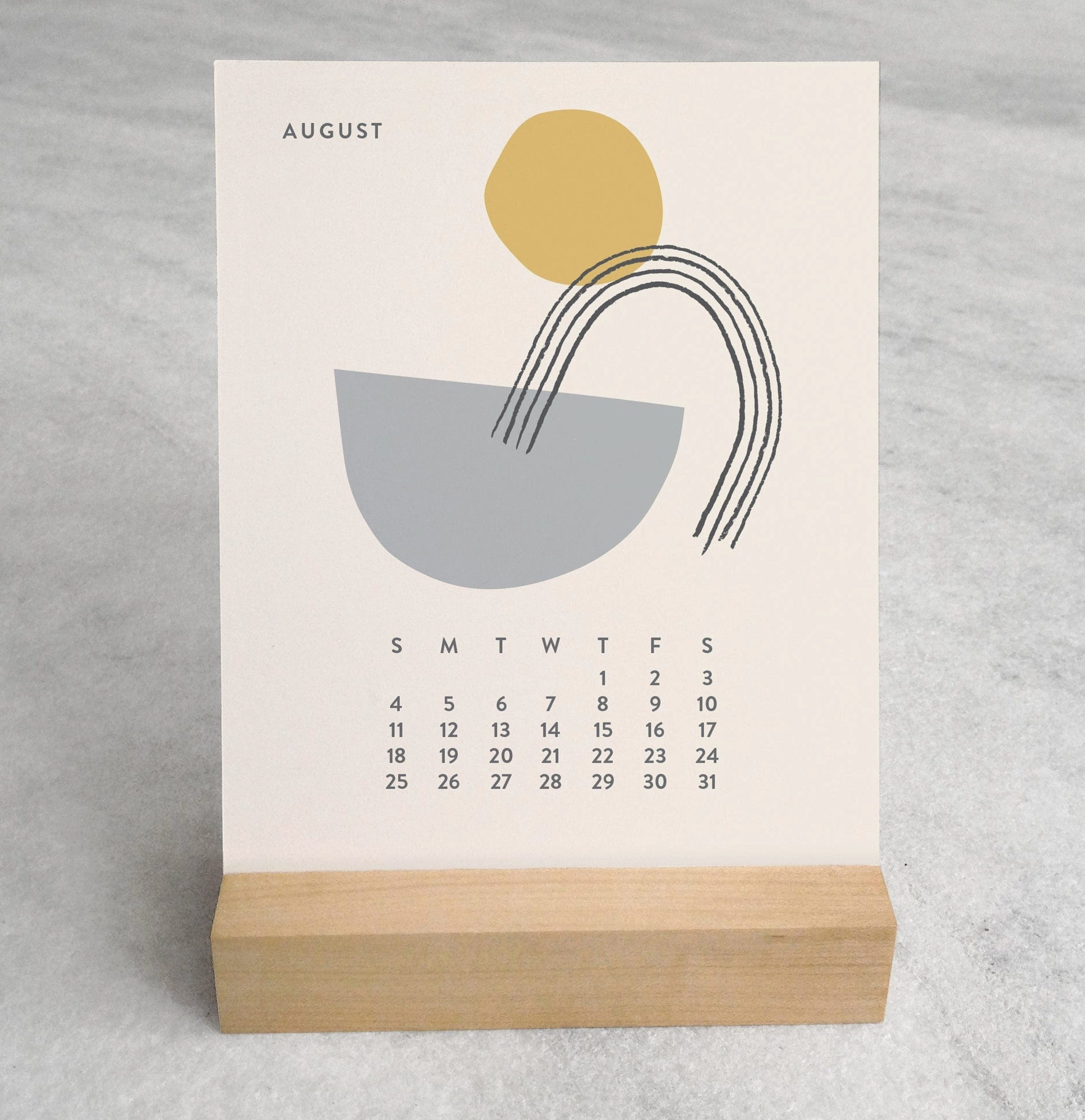 Close-up of the 2025 Desk Calendar August month with a unique monthly design visible on a 4.25" x 5.5" card. The calendar is positioned on a sturdy maple wood stand, emphasizing the quality craftsmanship. The calendar’s compact size is evident as it sits neatly on a small office desk.