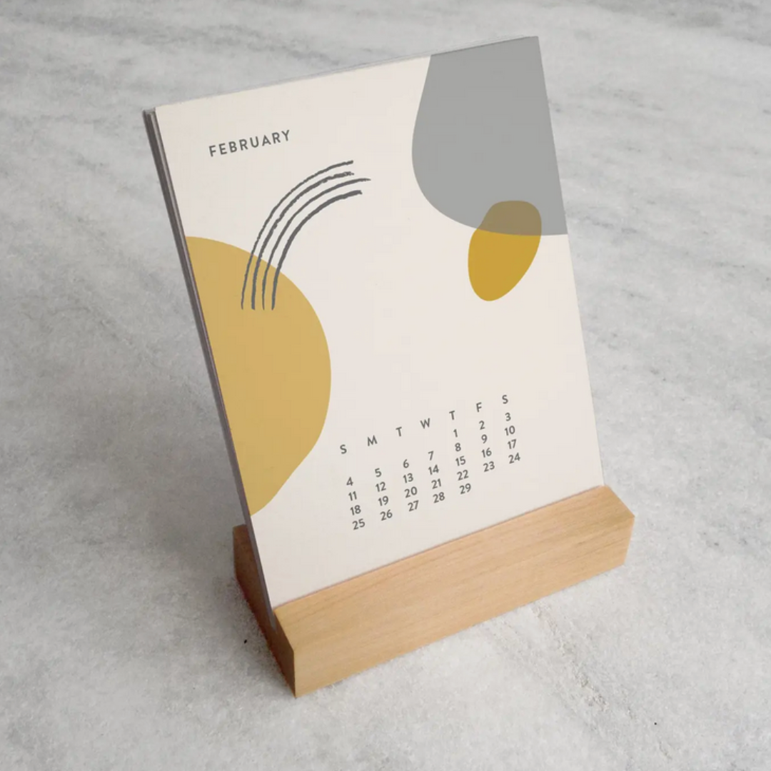 The 2025 Desk Calendar from Me To You Box is shown on a desk alongside a coffee cup and stationery. The calendar’s compact size and elegant design are highlighted, with the sturdy maple wood stand and unique monthly card designs adding a touch of charm to the workspace.