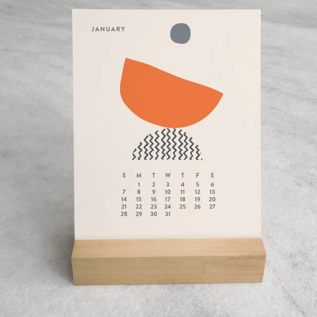 A side view of the 2025 Desk Calendar on a desk, showing the calendar's 4.25" x 5.5" card design and the sleek maple wood stand. The calendar is open to a page with intricate monthly artwork, blending well with a minimalist workspace.