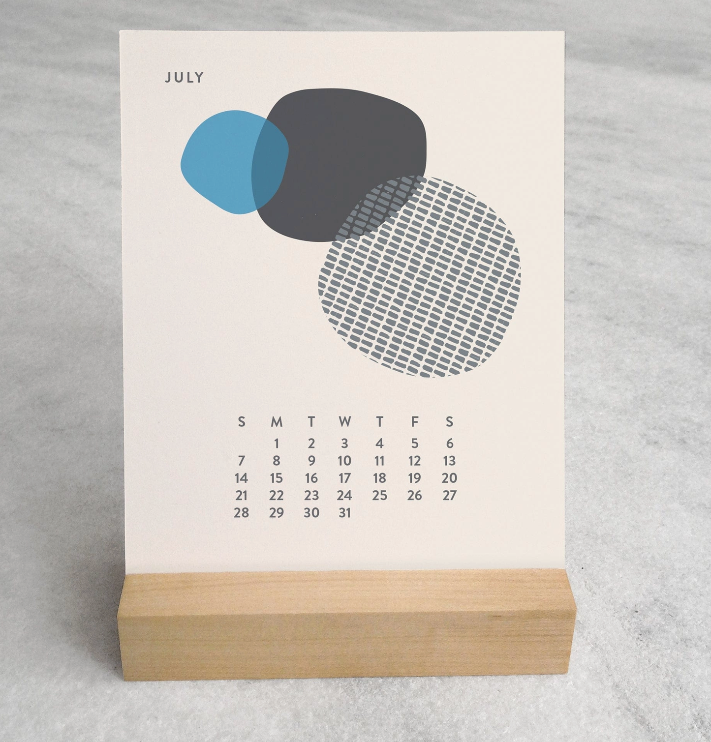 Close-up of the 2025 Desk Calendar July month with a unique monthly design visible on a 4.25" x 5.5" card. The calendar is positioned on a sturdy maple wood stand, emphasizing the quality craftsmanship. The calendar’s compact size is evident as it sits neatly on a small office desk.