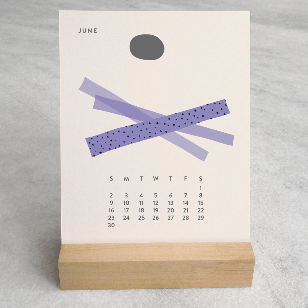 Close-up of the 2025 Desk Calendar June month with a unique monthly design visible on a 4.25" x 5.5" card. The calendar is positioned on a sturdy maple wood stand, emphasizing the quality craftsmanship. The calendar’s compact size is evident as it sits neatly on a small office desk.