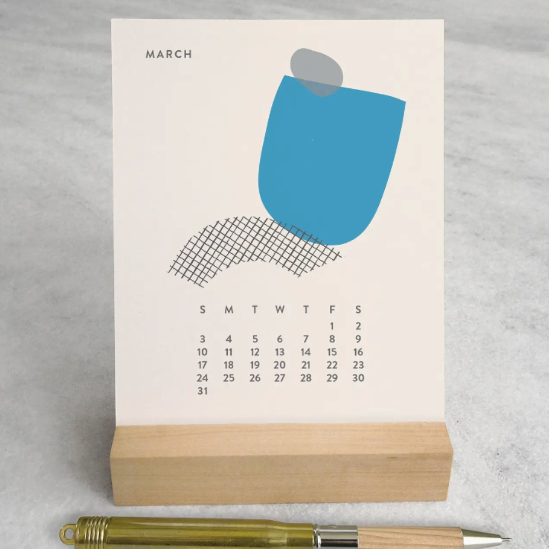 A top-down view of the 2025 Desk Calendar on a desk, revealing the sturdy maple wood stand and the calendar's 4.25" x 5.5" cards. Each card displays a different monthly design, adding visual interest and practicality to the small desk area.