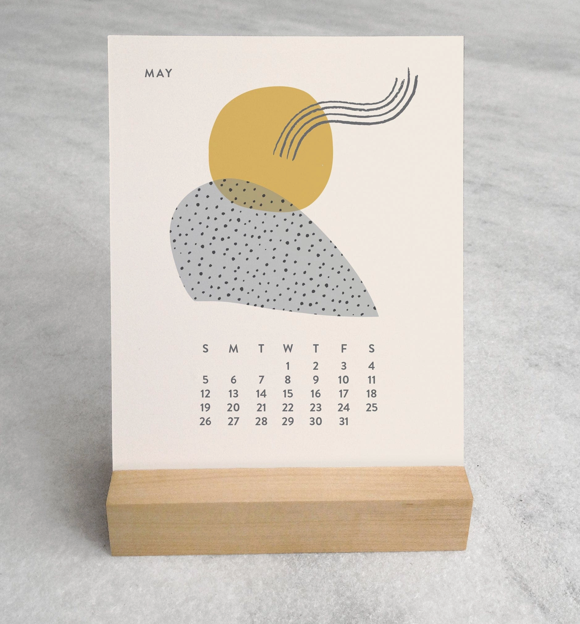 The 2025 Desk Calendar displayed on a table. The calendar is set on a desk with a focus on its maple wood stand and the monthly design cards. The compact size and elegant presentation are showcased, making it clear that this calendar is both a functional and stylish addition to any workspace.