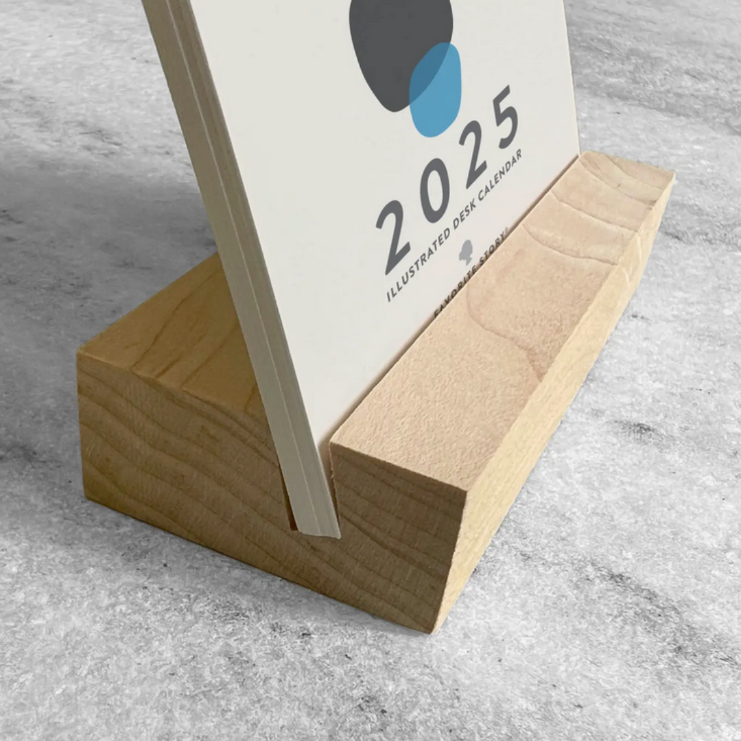 2025 Desk Calendar by Me To You Box displaying a beautifully designed front card on a sleek maple wood stand. This compact 4.25" x 5.5" calendar offers unique monthly artwork, bringing inspiration and organization to any desk space. Made in the USA.