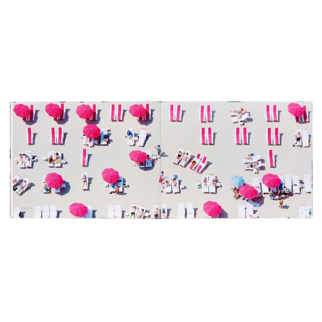 An open spread of a sandy beach. The photo is taken with a camera from a helicopter over the beach. The beach is filled with people lounging with vibrant pink colored beach towels and pink beach umbrellas.