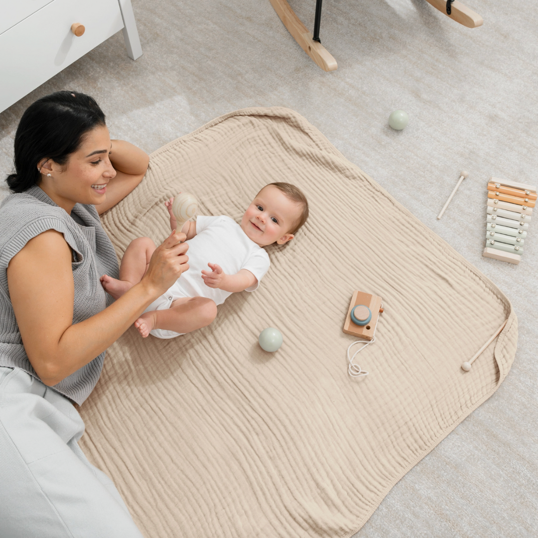 Woman and baby on a beige blanket with wooden toys in a nursery.