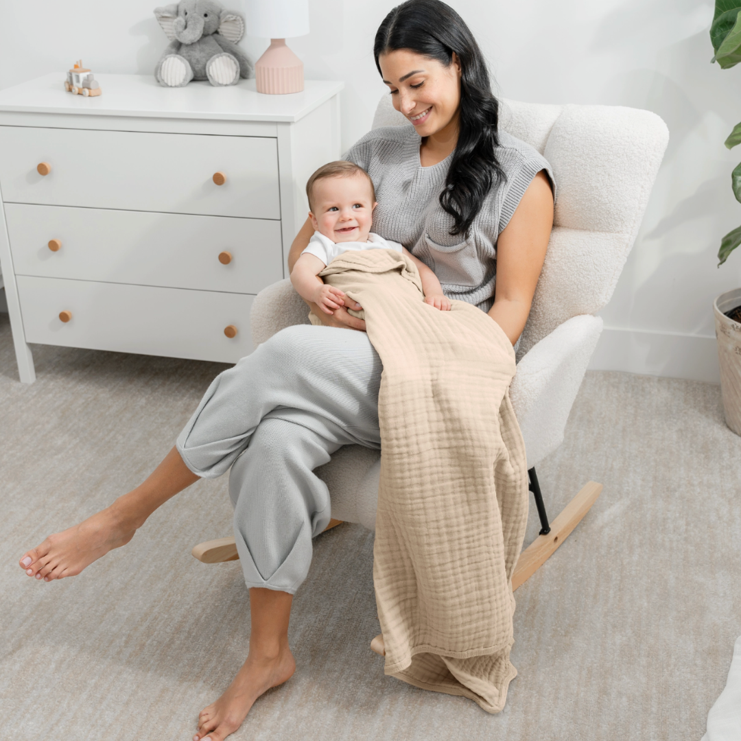 Woman sitting in a rocking chair holding a baby with a beige blanket, in a nursery setting.