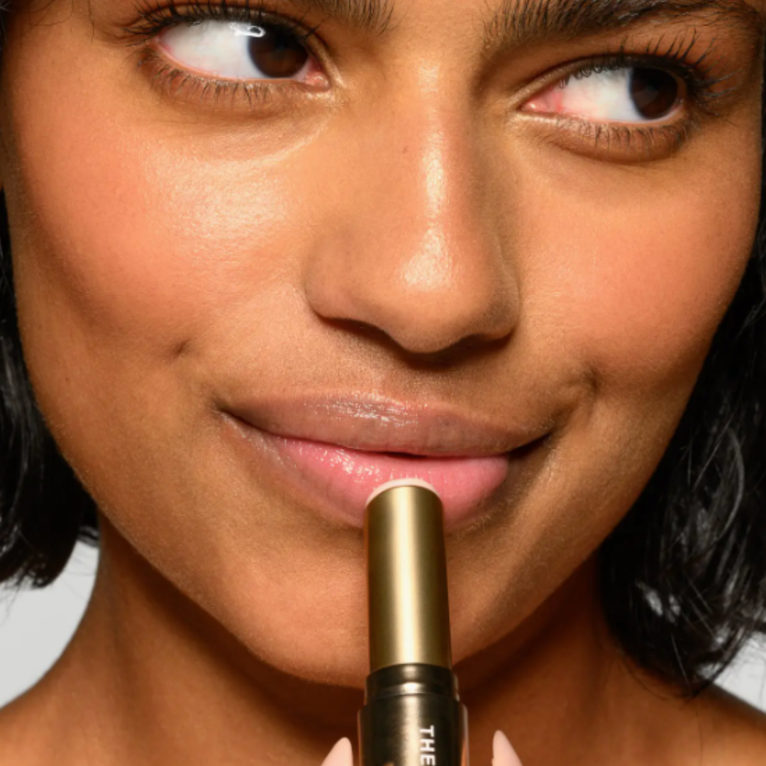 Close-up of a woman holding a gold lipstick tube against a neutral background.
