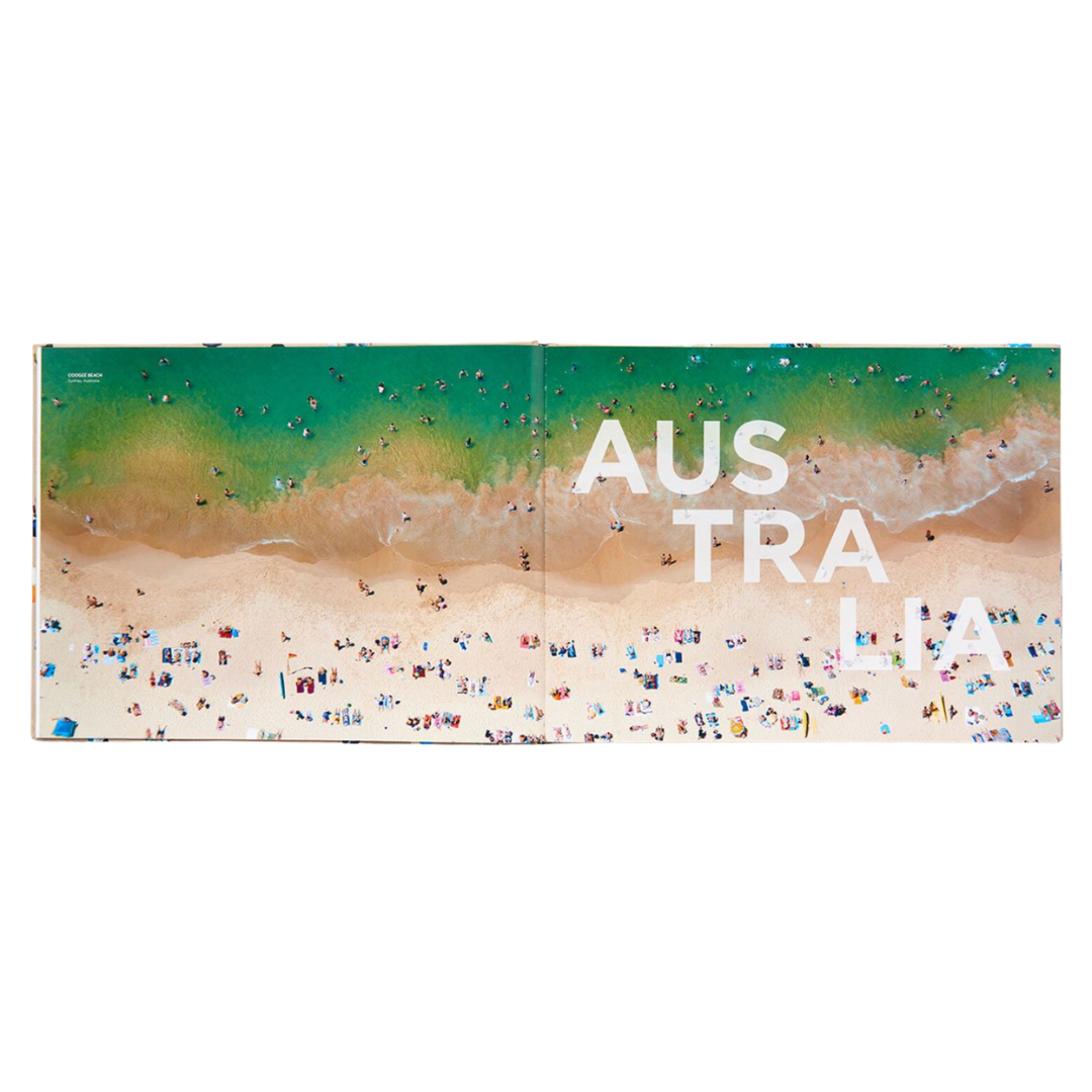 An open spread of a beach in Australia. The photo is taken with a camera from a helicopter over the beach. The beach is filled with people swimming and lounging with vibrant colored beach towels and beach umbrellas.