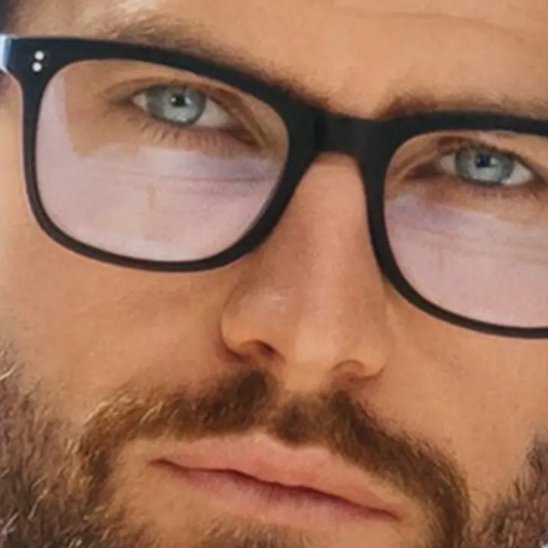 Close-up of a man with a beard and blue eyes wearing black rectangular eyeglasses with light pink-tinted lenses.