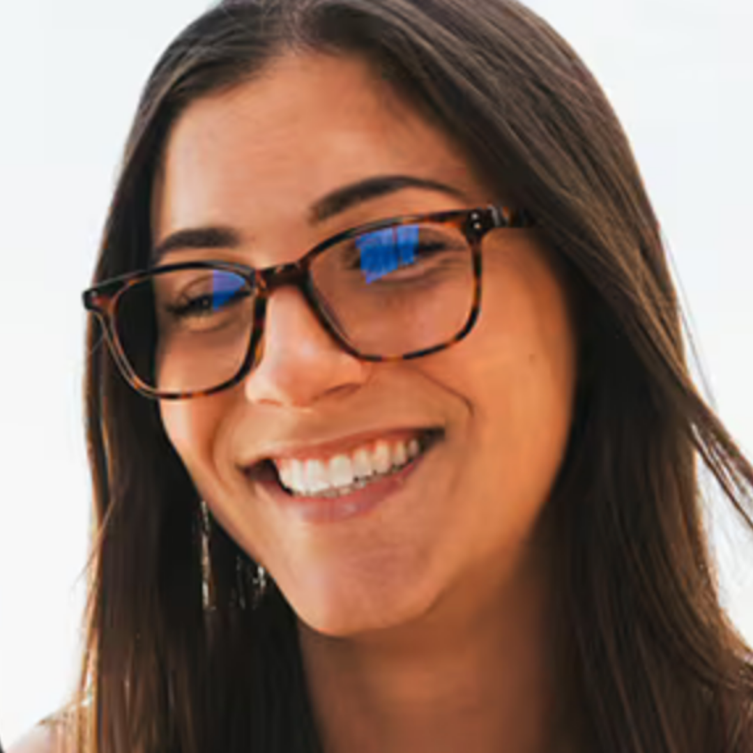 A close-up portrait of a smiling woman with long dark brown hair wearing tortoiseshell eyeglasses against a bright white background.