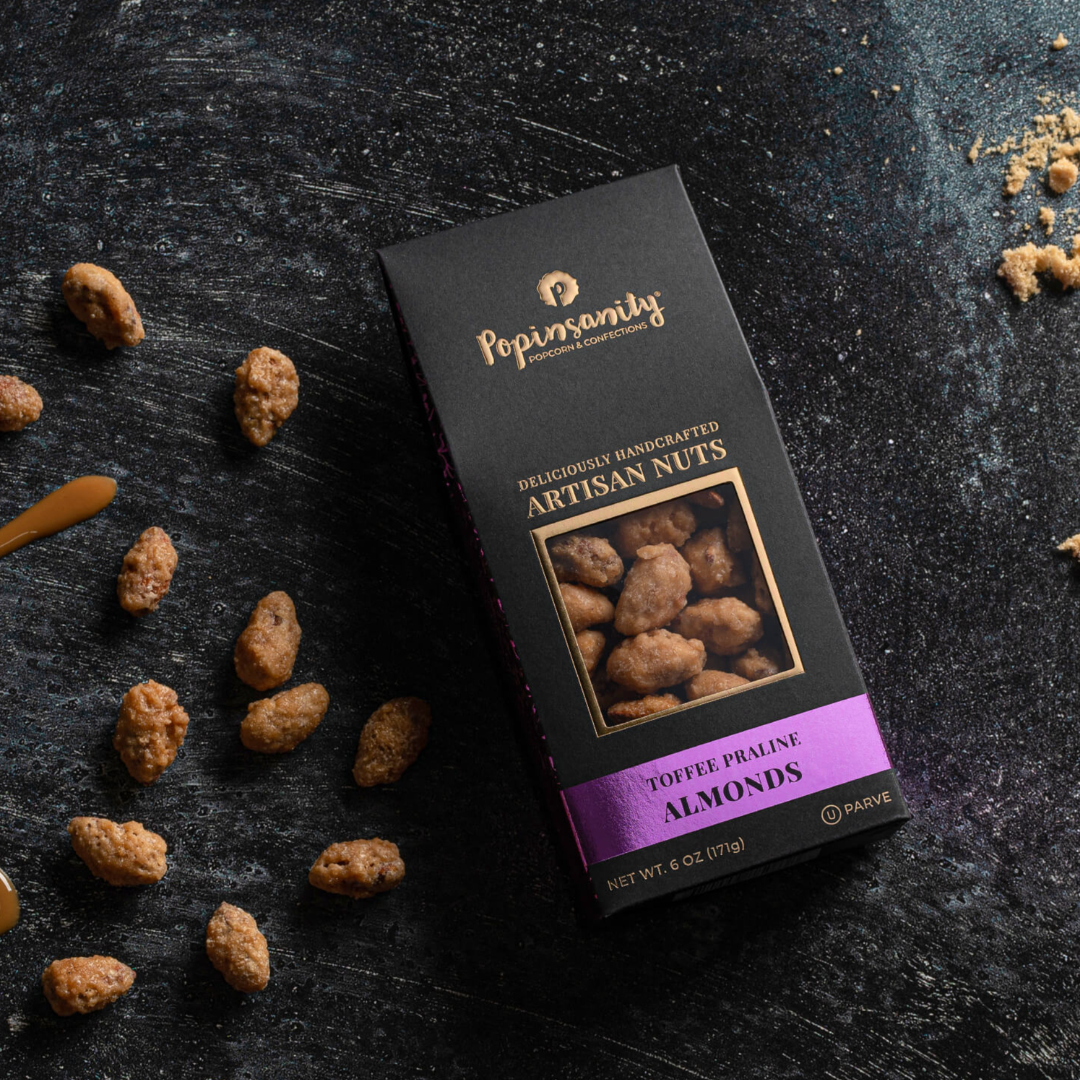 Box of Popinsanity Artisan Nuts on a dark surface with almonds around.