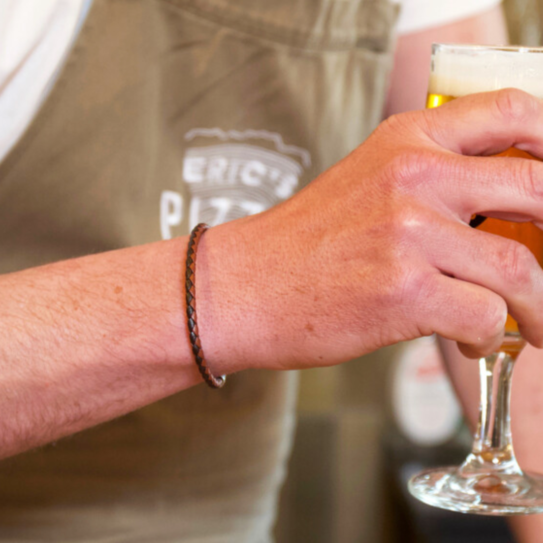 Brown slim leather bracelet shown on a man's wrist. The man is holding a beer in a clear glass with foam on top.