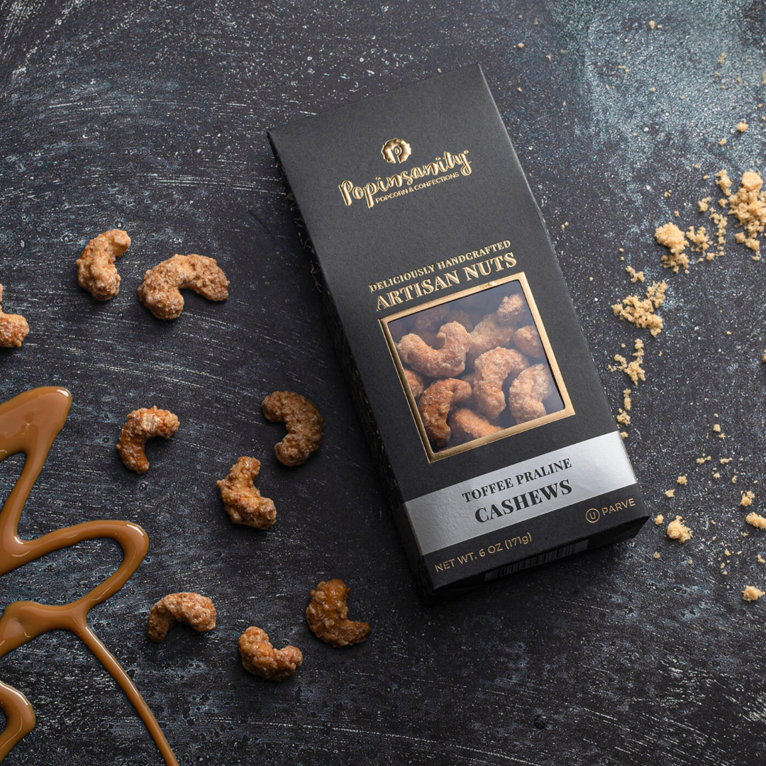 Box of Poppin' Good toffee praline cashews on a dark surface with nuts and caramel.