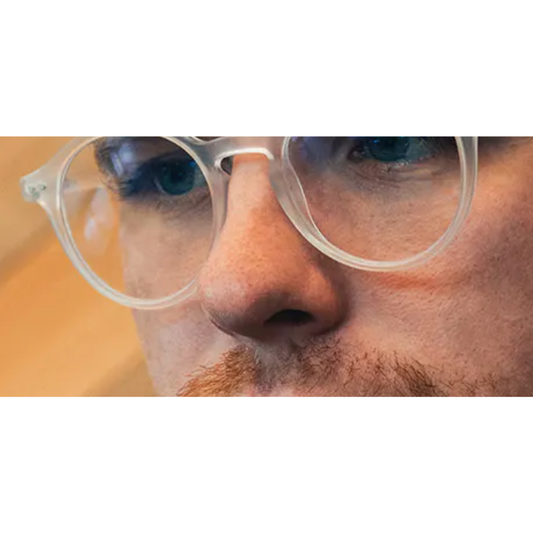 Close-up of a person wearing clear round eyeglass frames, with the nose and part of the eyes visible against a warm beige background.