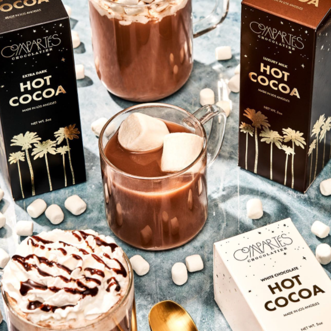 A five-ounce package of Compartés Luxury Hot Cocoa Mix, featuring award-winning drinking chocolate, elegantly wrapped with a stunning LA sunset palm silhouette design and reflective golden foil, perfect for indulging in a rich, milky hot chocolate experience. Image also shows mug of cocoa with marshmallows floating on top.