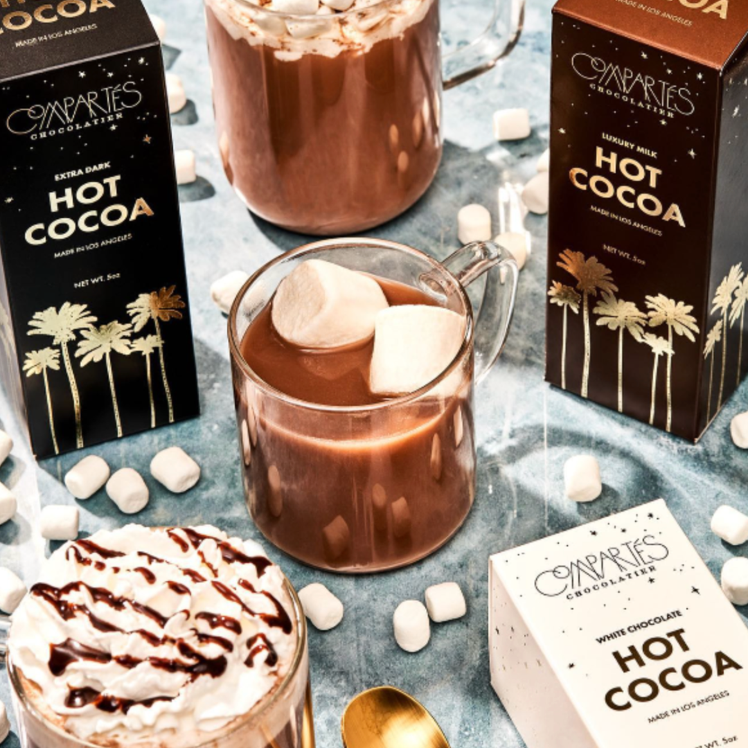 Hot cocoa with marshmallows, Cooperties chocolate bars, and a spoon on a textured surface.