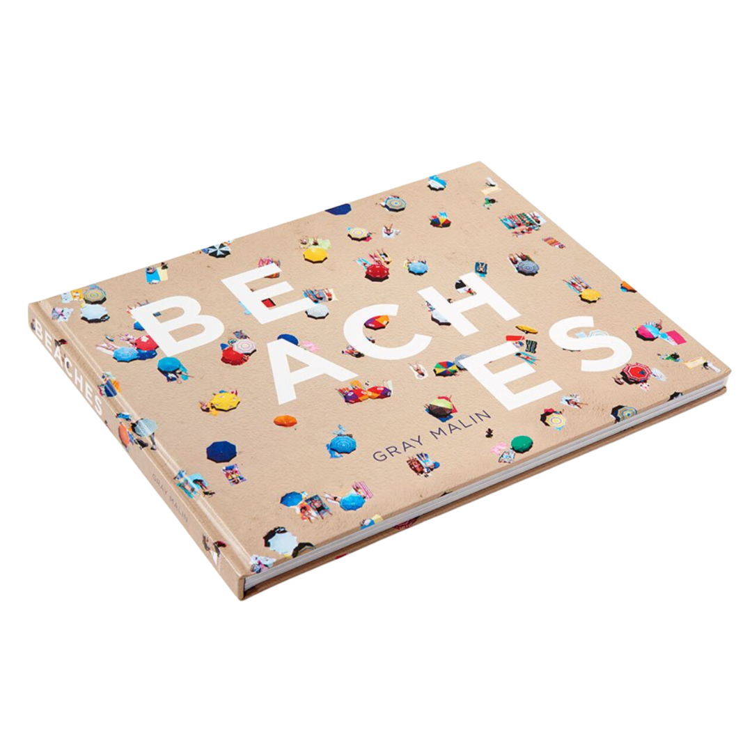 Cover of Beaches Coffee Table Book by Gray Malin displayed on an angle with white background. The cover shows a beach with different colored sun umbrellas.  The cover's photo is taking from the top down.