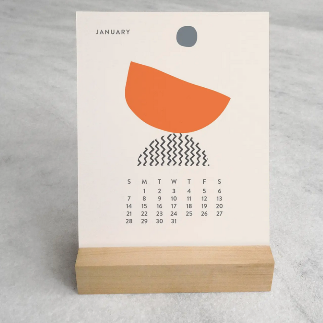 2025 Desk Calendar by Me To You Box displaying a beautifully designed 'January' card on a sleek maple wood stand. This compact 4.25" x 5.5" calendar offers unique monthly artwork, bringing inspiration and organization to any desk space. Made in the USA.