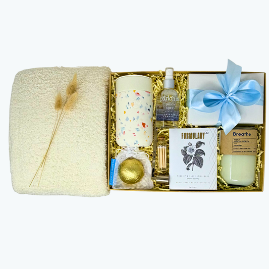A gift box with cozy blanket, cream silicone and ceramic tumbler, facial mask, candle, matches, shower steamer and pillow mist on a white background.