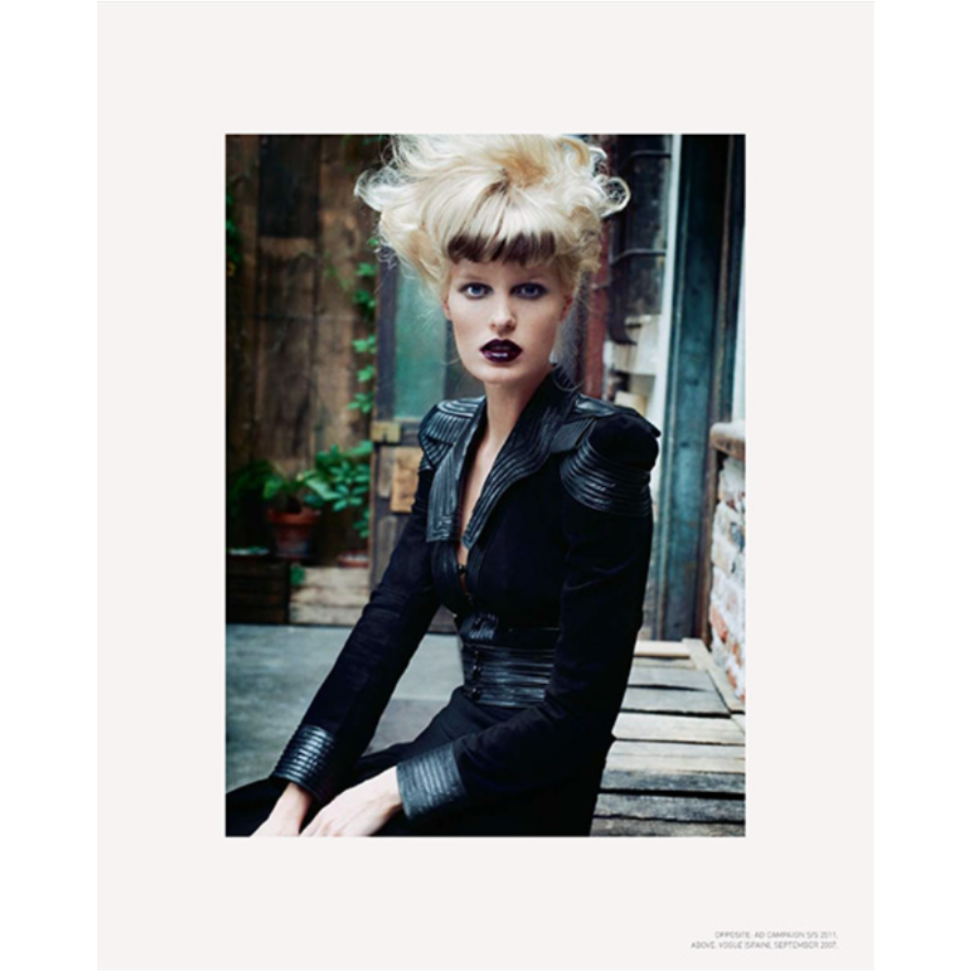 Female Gucci model dresses in black with blond hair in a messy bun on top of her head.  The model has very dark lipstick on and is sitting outside on a wooden plank bench.