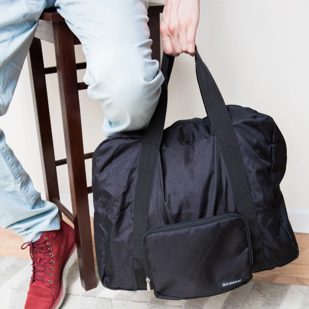 Man holding a black fold able duffel bag in his left hand. The duffel bag is large enough for weekend getaways and has a front outside pocket. Versatile and practical and light weight.