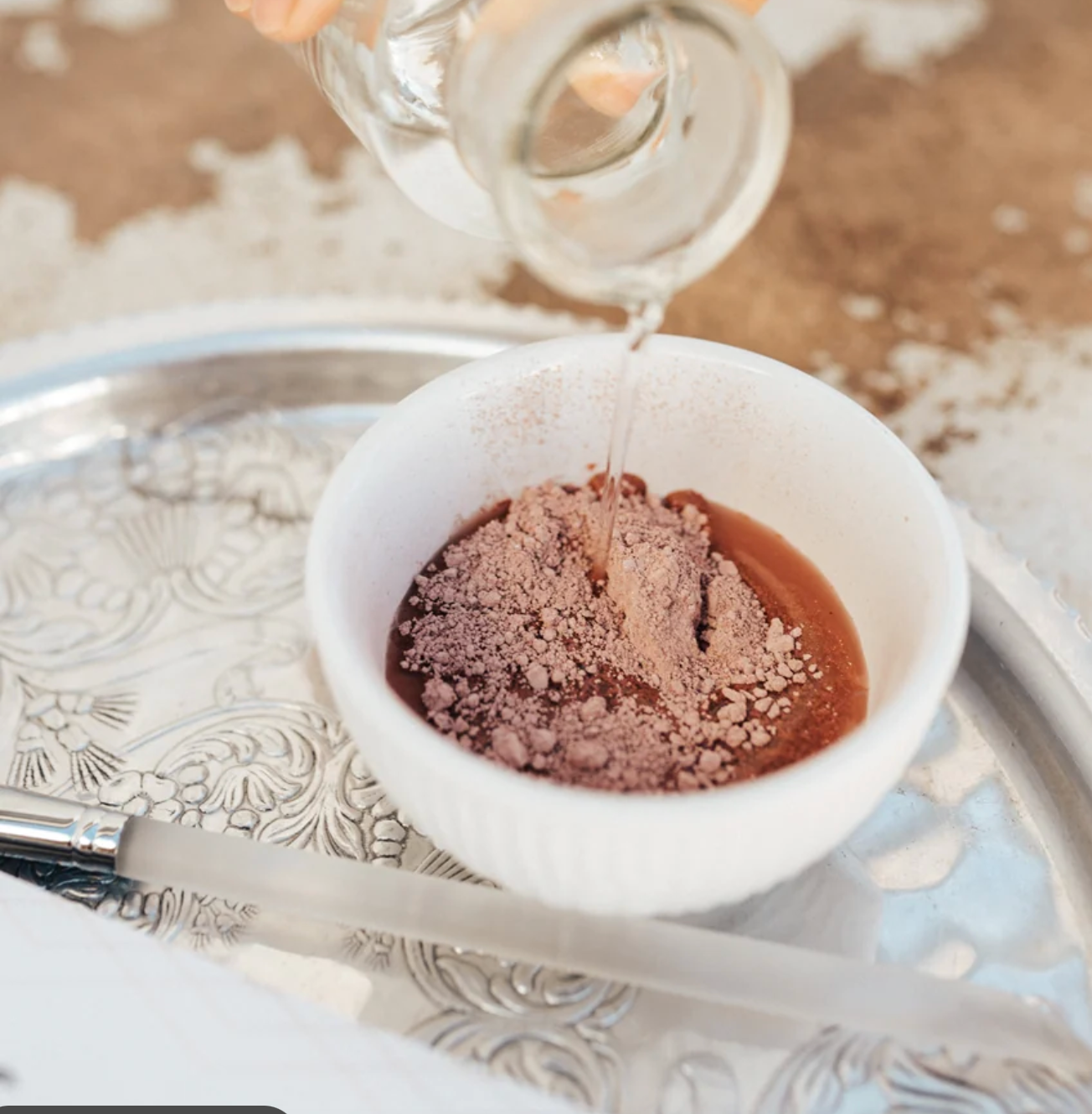 Formulary 55 facial mask pink clay powder in a white bowl with water being added to make a face mask mix. The bowl is set on a silver tray.