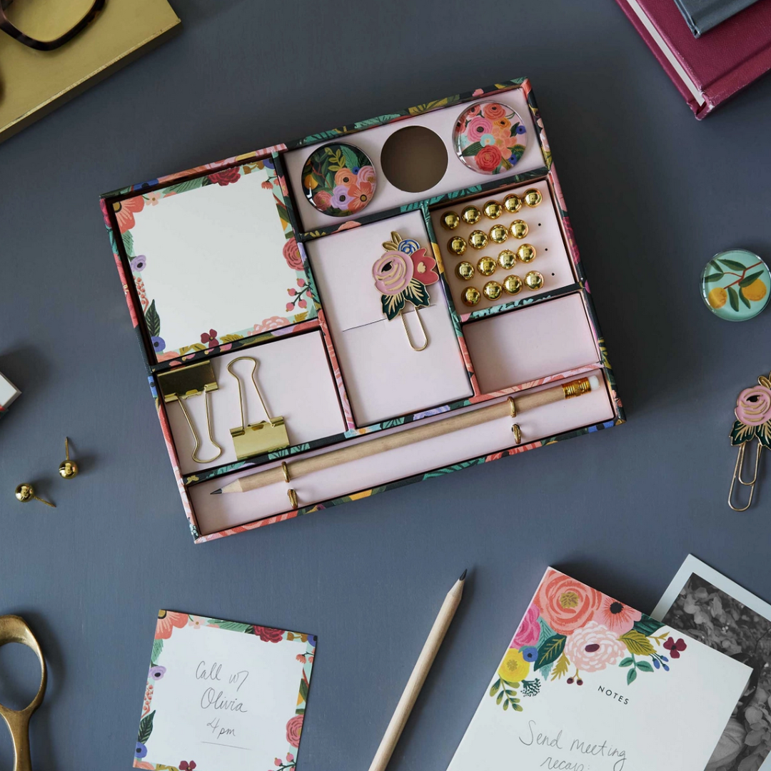 Colorful floral-themed box containing office supplies. Includes a tear-off pad, magnets, enamel paper clips, gold push pins, eraser, binder clips, and pencils.