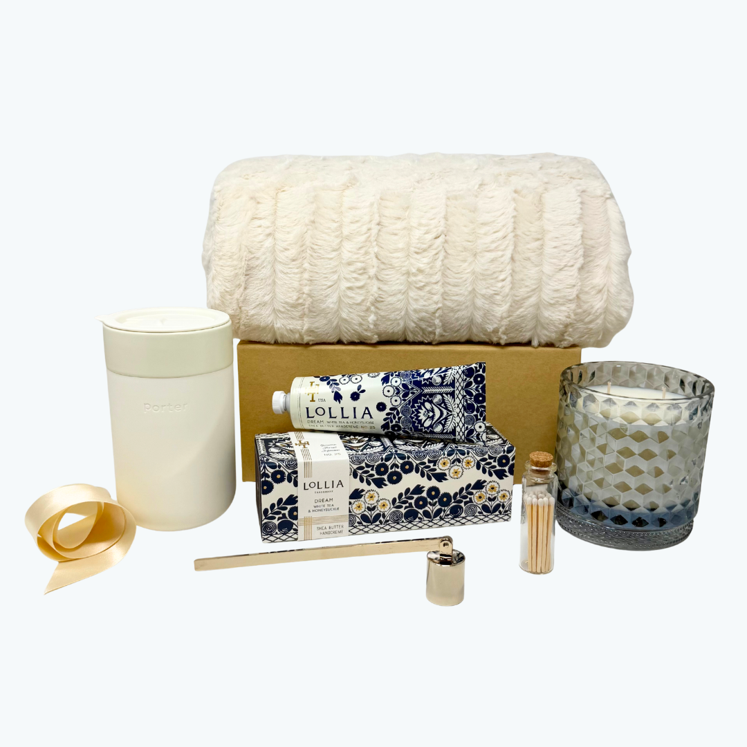 A curated self-care and relaxation gift set arranged on a light gray background. The set includes a cream-colored textured blanket, a grey glass candle jar, a patterned Lollia hand cream tube, Lollia hand cream box, a gold candle snuffer, cream silicone wrapped 16 oz mug and a small bottle of matches.