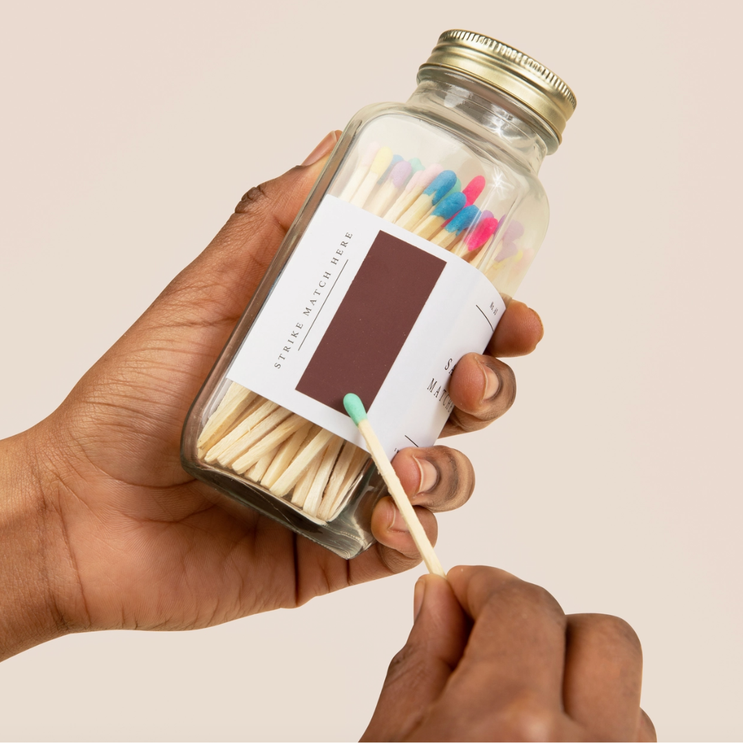 Hand holding a glass jar of matches with colorful heads against a beige background. A person's hand is striking a light green tipped match against the strike pad on the bottle.