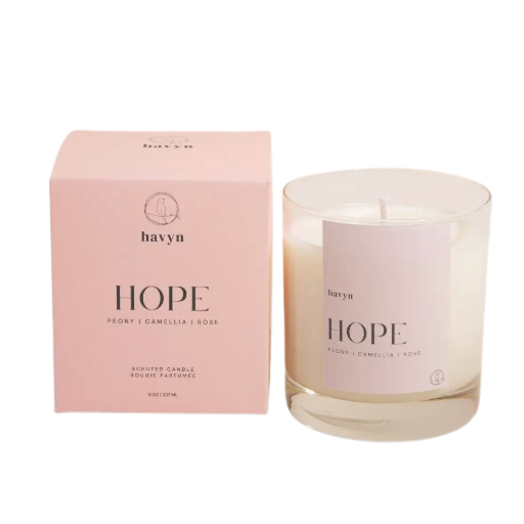 A close-up of the HOPE Signature Candle, featuring an elegant glass vessel with the word "HOPE" prominently displayed. The candle is made from a 100% coconut and soy wax blend and is hand-poured in San Diego. It features a serene blend of Peony, Camellia, Rose, Pomelo, and Mandarin scents, symbolizing hope and tranquility. The candle's clean fragrance is free of allergens and carcinogens, and it is designed to burn for up to 40 hours.