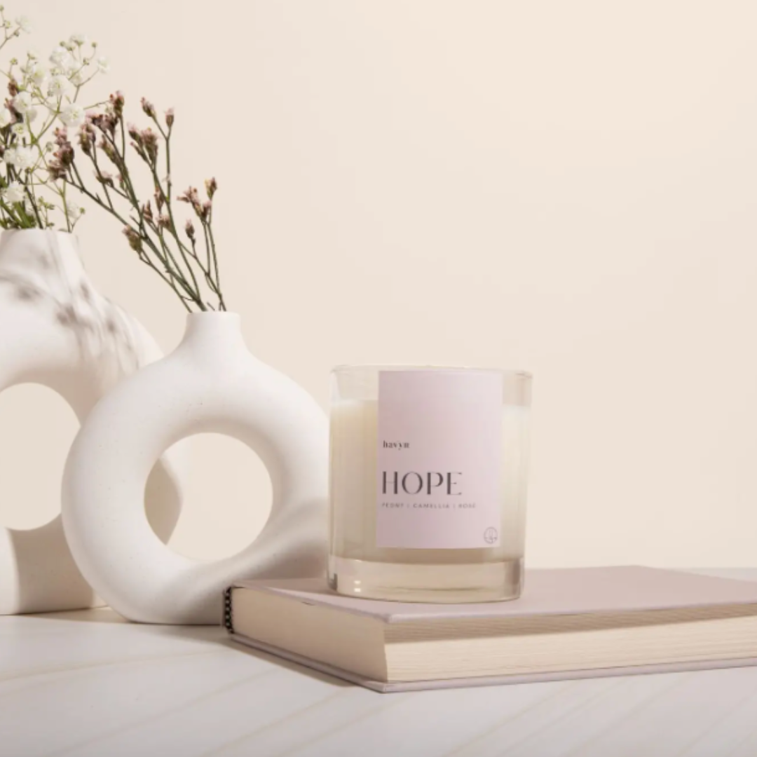 A view of the HOPE Signature Candle placed on a soft, neutral background. The candle is housed in a transparent glass vessel marked with the word "HOPE." It is crafted from a blend of coconut and soy wax and features a soothing fragrance of Peony, Camellia, Rose, Pomelo, and Mandarin. The candle is hand-poured in San Diego and offers a long burn time of 40 hours, intended to provide comfort and encouragement.