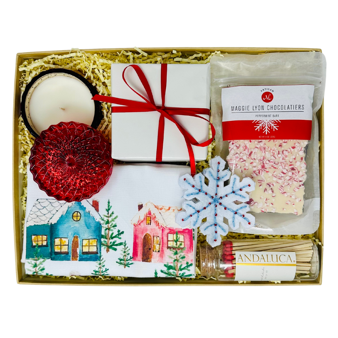 The "HAPPY HOLIDAYS" Me To You Box is shown, packed with festive items including a red beaded snowflake ornament, an 8-ounce bag of Maggie Lyon Peppermint Bark, a colorful winter cottages kitchen towel, a red glass soy wax candle with holiday spice scent, and a glass jar of matches with a cork top. Each item is arranged to reflect the holiday spirit and the thoughtful curation, with a note about the charitable contribution supporting children’s care programs. The box promises a delightful holiday experience