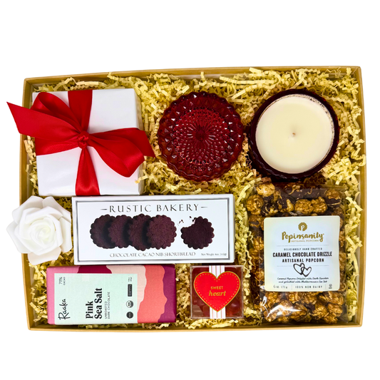 A valentine's gift displayed inside a box with a red glass shimmer candle, organic pink sea salt dark chocolate bar, white box of chocolate shortbread cookies, strawberry gummy hearts in an acrylic cube and a clear bag of caramel chocolate drizzle artisanal popcorn. The gift is wrapped in white gloss paper with a red satin bow.