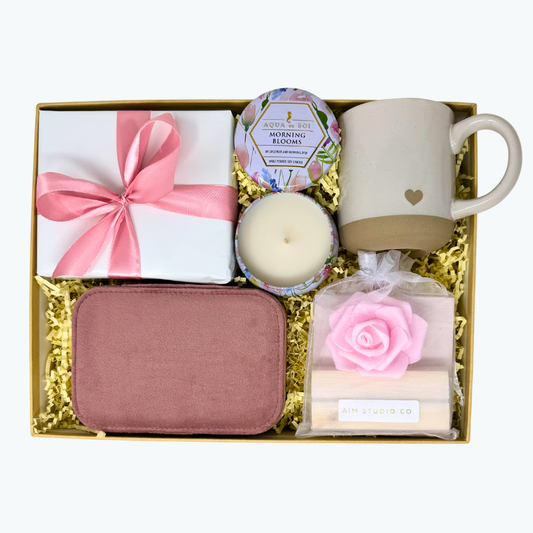 Gift set with heart stoneware mug, morning blooms tin candle with flowers, set of positive affirmation cards with wooden stand, and pink velvet jewelry box all displayed in a box.