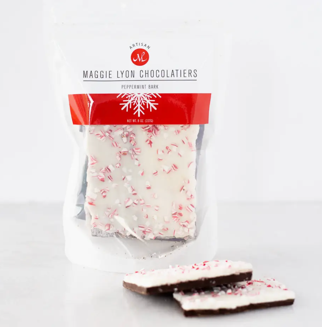 An 8-ounce bag of Maggie Lyon Chocolatiers Peppermint Bark, featuring layers of rich dark chocolate and creamy white chocolate, topped with crunchy peppermint pieces. The festive packaging highlights the gourmet quality and seasonal appeal of this holiday treat, ideal for gifting or personal enjoyment.