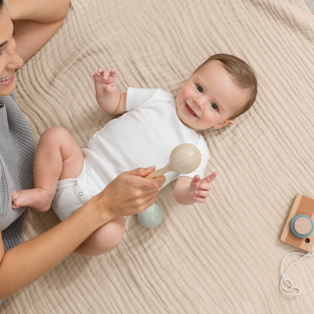 Baby lying on a beige muslin blanket with a woman holding a wooden rattle.