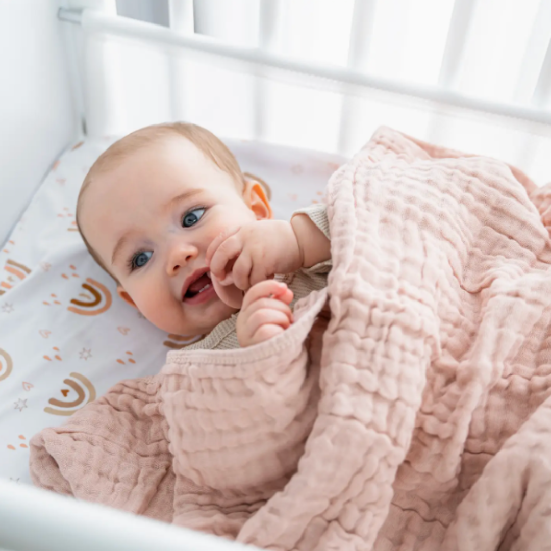 Baby wrapped in a pink muslin blanket lying in a crib with a white background.