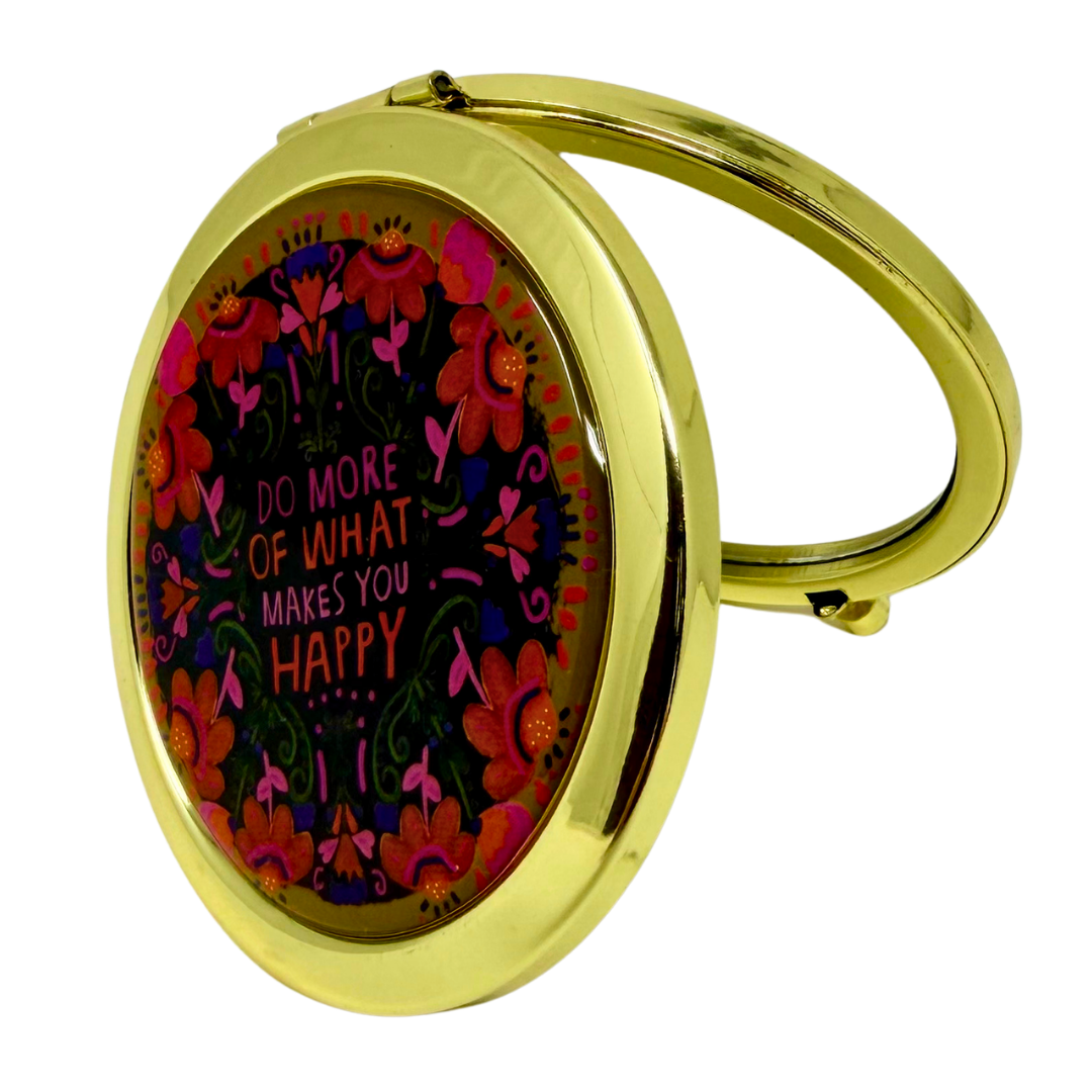A gold oval compact mirror with a hinged lid, featuring a colorful floral design and the quote 'DO MORE OF WHAT MAKES YOU HAPPY' on the front, set against a plain white background.