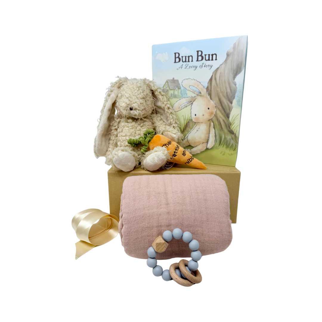 Baby gift set with plush bunny, plush orange carrot, pink blanket, book and teething ring on a white background.