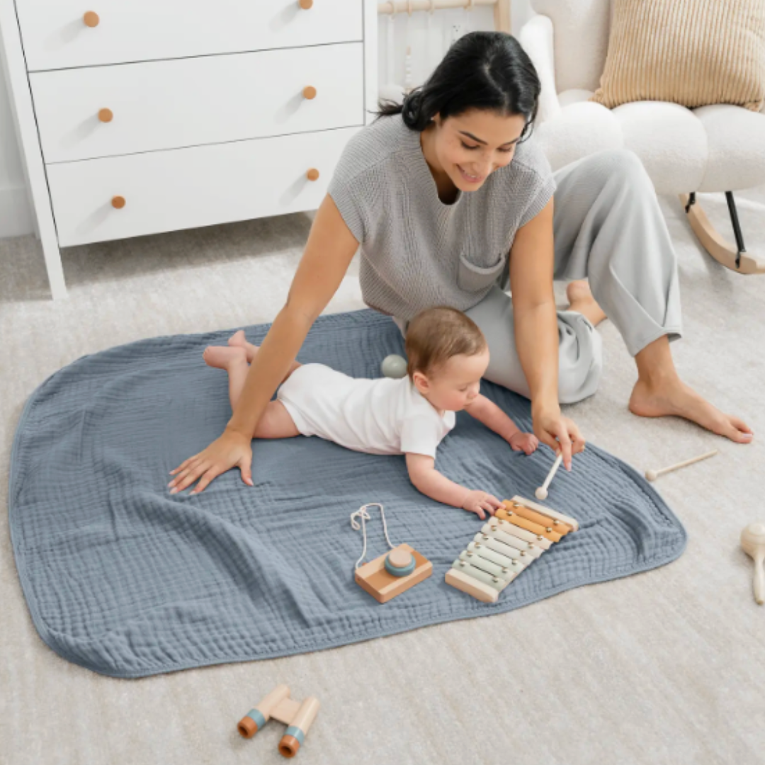 Mom and baby on a blue muslin blanket in a nursery with toys.