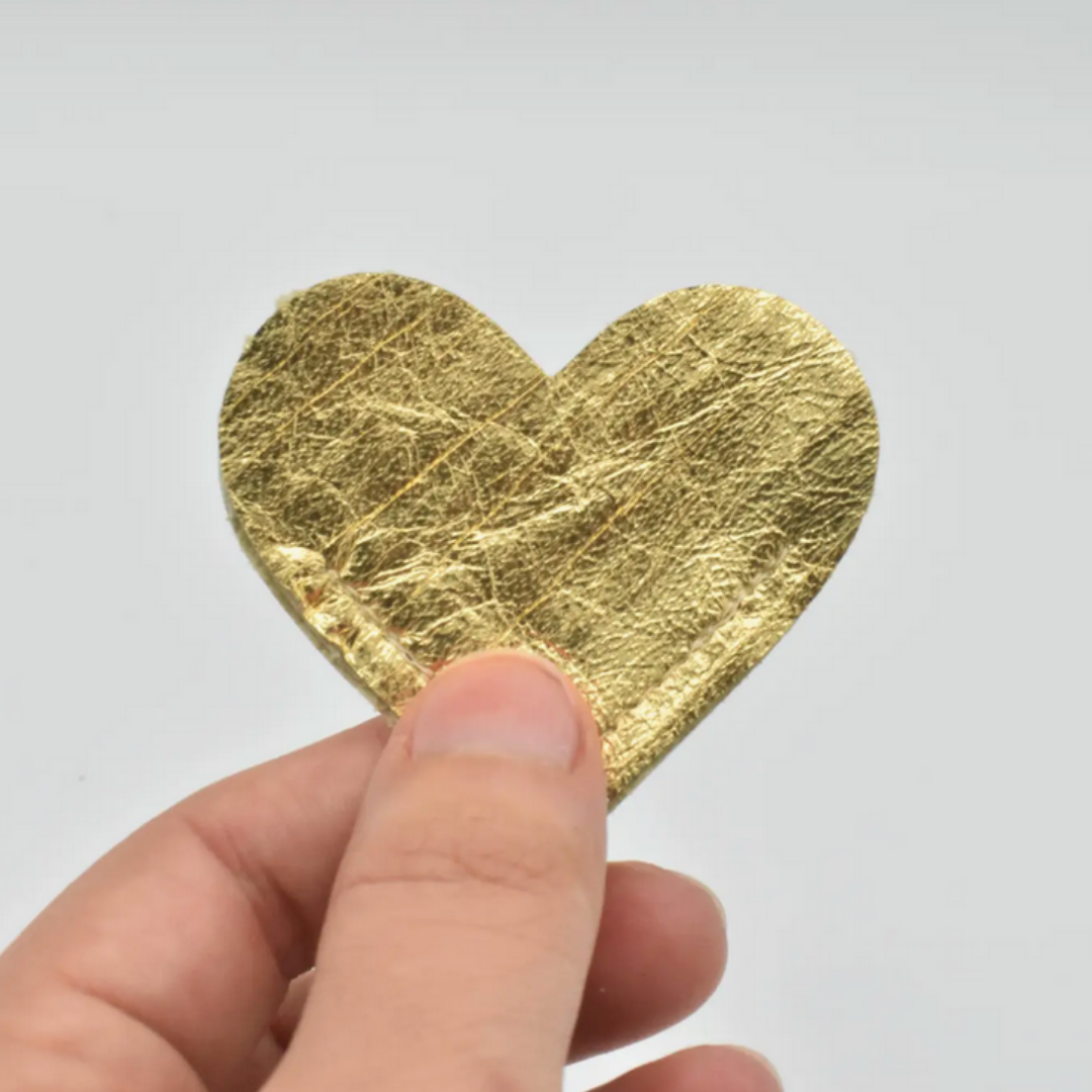 A hand holding a small, textured gold heart-shaped piece against a plain light gray background.