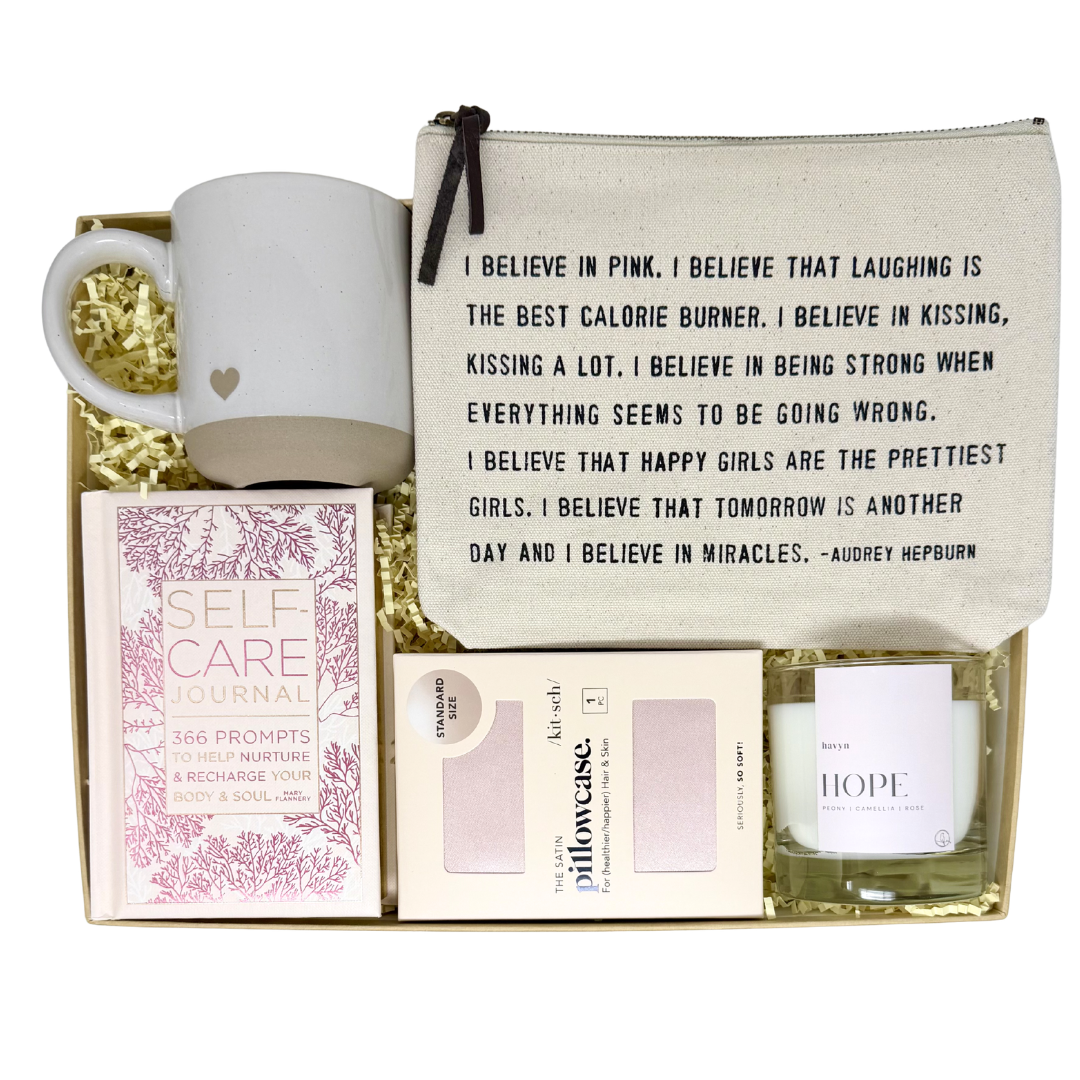 Curated gift neatly displayed on top of crinkle paper in a box on a white background. Included is a stoneware mug with a heart on it, pink satin pillowcase, pink self care journal, "hope" glass candle and canvas pouch with an Audrey Hepburn quote.