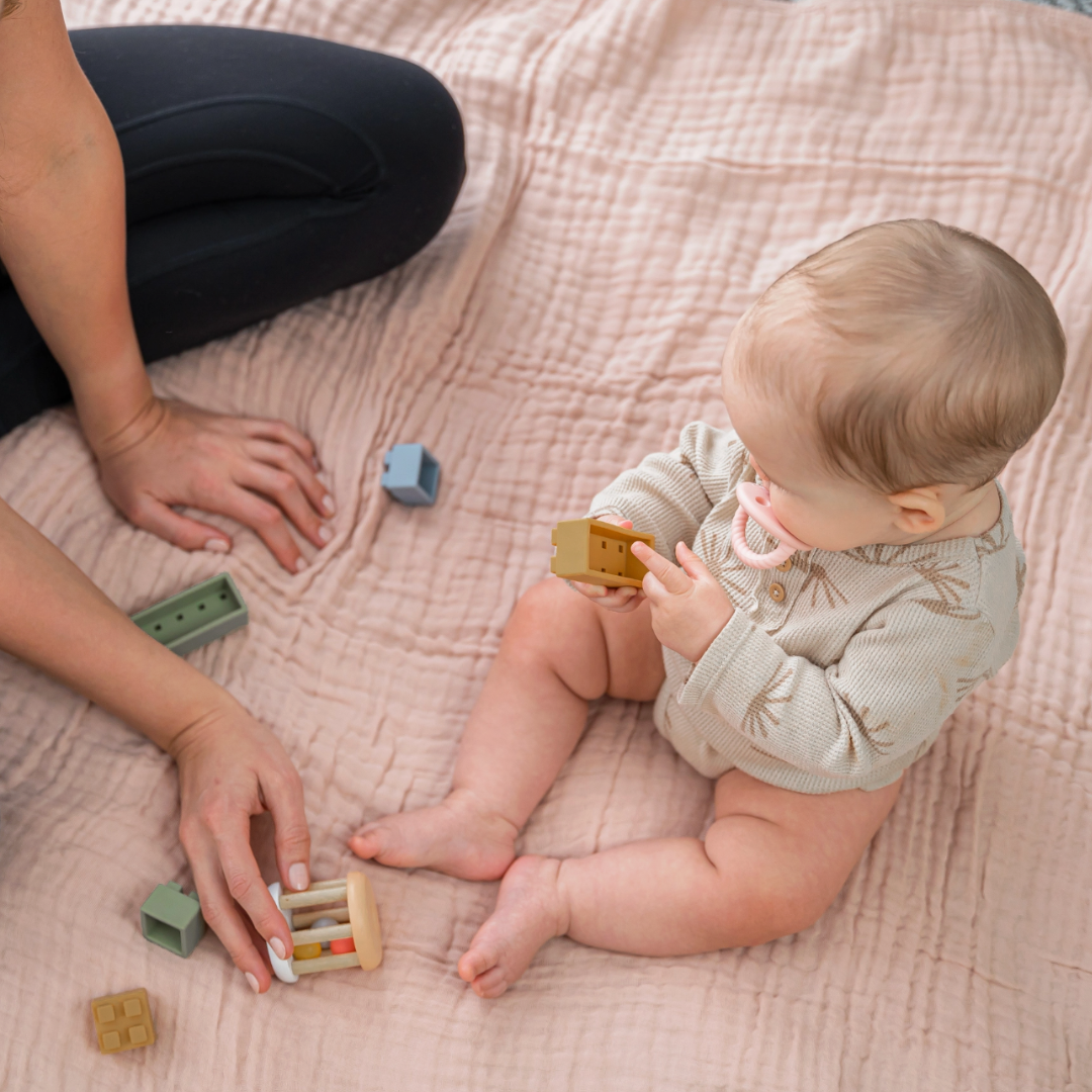 Baby playing with wooden toys on a pink muslin blanket with an adult's hands nearby.