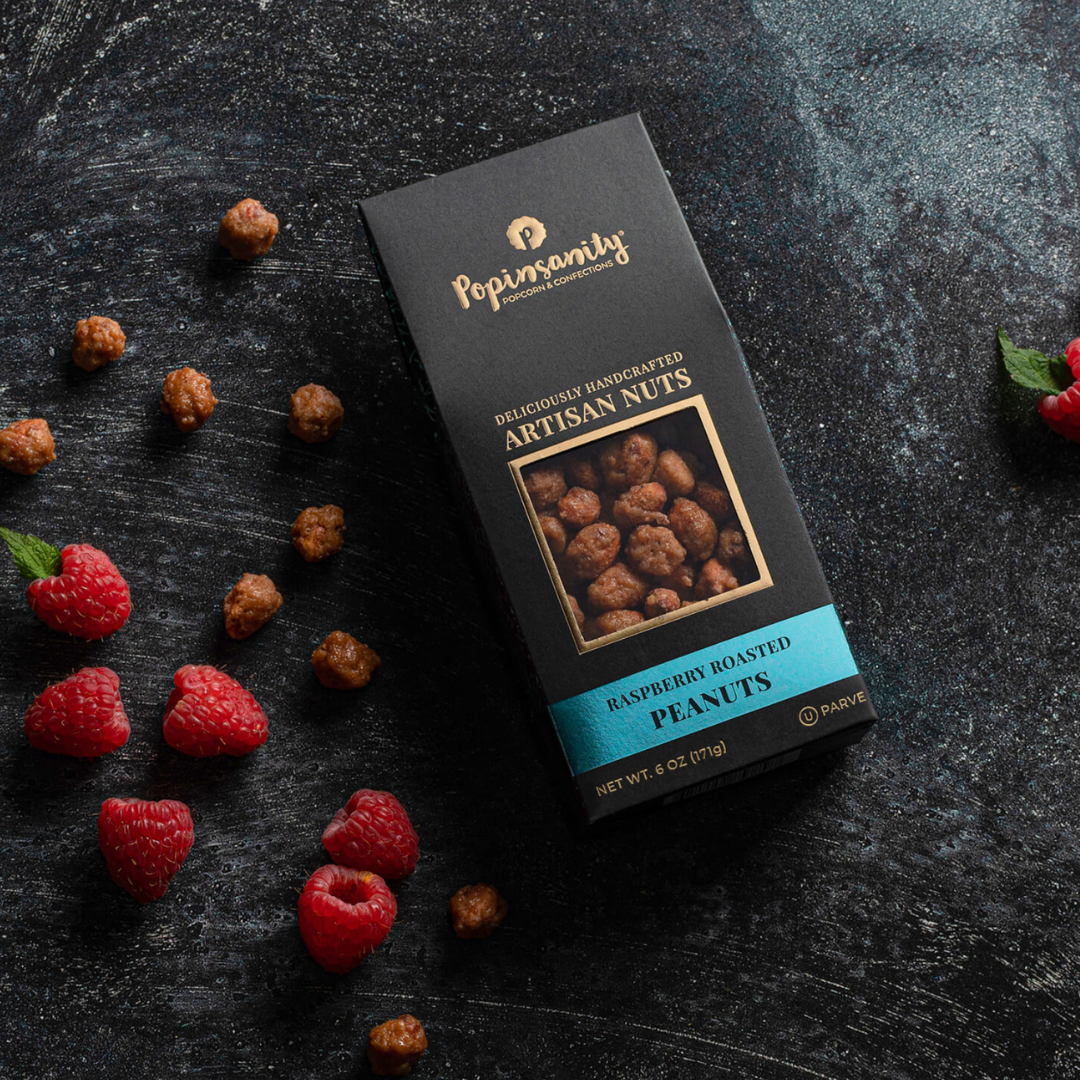 Box of Popin' Nuts Raspberry Roasted Peanuts on a dark surface with raspberries.